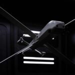 A futuristic HX-2 drone with a sleek, gray body and long wings, suspended in a dark, high-tech hangar with glowing lights.