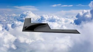 B-21 Raider stealth bomber flying above cloud layer in a clear blue sky, viewed from above.