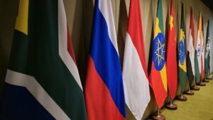 A row of international flags on wooden stands against a yellow wall, including the flags of Russia, China, Ethiopia, Brazil, India, and other BRICS member and partner nations displayed at a diplomatic summit.