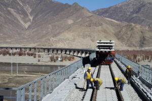 Workers in yellow helmets build railway tracks on a bridge in Tibet, surrounded by mountains and dry terrain.