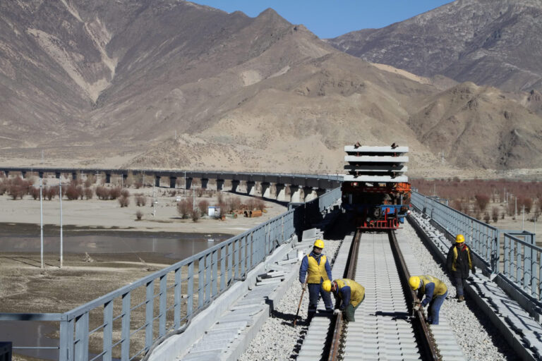 Workers in yellow helmets build railway tracks on a bridge in Tibet, surrounded by mountains and dry terrain.