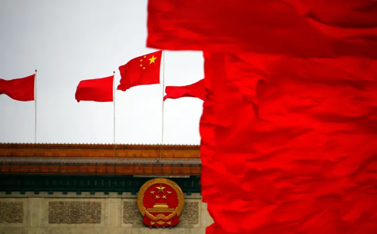 A red Chinese flag with yellow stars waving on a pole, set against the backdrop of a government building and Tiananmen Square.
