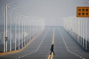 A lone person in a dark coat walks down the middle of an empty highway in China, surrounded by evenly spaced streetlights and a foggy atmosphere. A sign with Chinese characters is visible on the right side of the road.