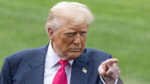 A close-up image of former President Donald Trump in a blue suit and red tie, pointing forward with a serious expression, standing against a green grass background with an American flag pin on his lapel.