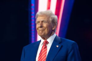 Donald Trump, dressed in a blue suit with a red and white striped tie, stands with a small American flag pin on his lapel, smiling against a colorful background of red, white, and blue lights.