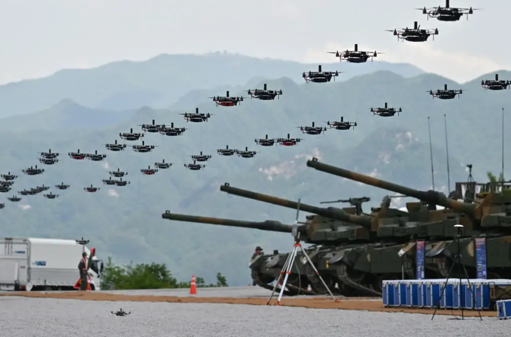 A large group of drones flying in an organized pattern above several military tanks stationed on a gravel field, with mountains in the background and a few personnel and vehicles present.