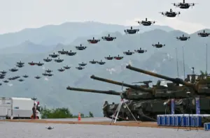 A large group of drones flying in an organized pattern above several military tanks stationed on a gravel field, with mountains in the background and a few personnel and vehicles present.