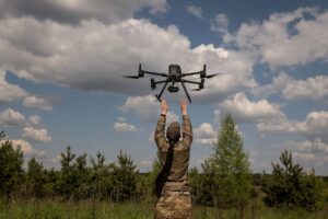 A soldier in camouflage raises their hands to launch a multi-rotor drone into a partly cloudy sky, surrounded by green fields and trees in a rural landscape.