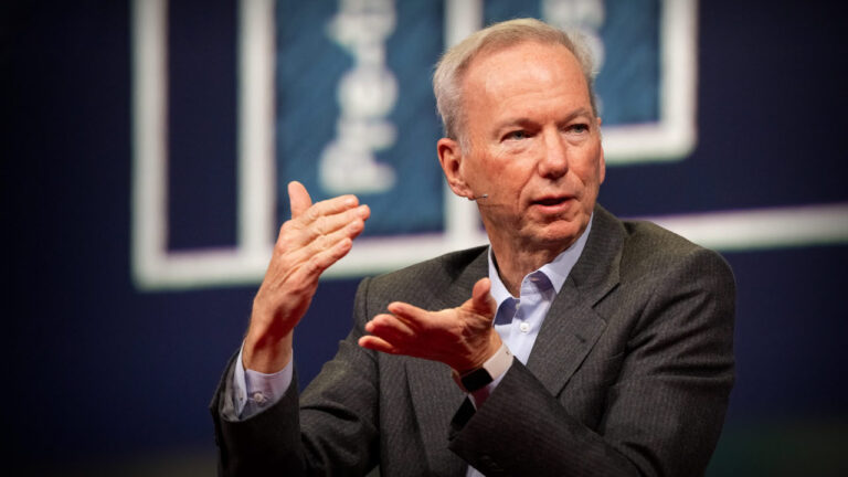 Eric Schmidt speaking on TED stage in gray suit and blue shirt, hands raised in gesture, with blue conference background.