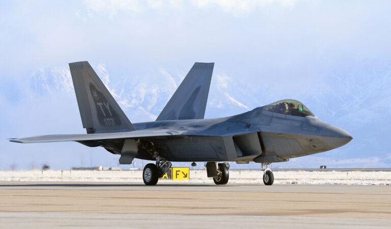 A gray F-22 Raptor fighter jet with "TY" markings on its tail taxis on a runway, surrounded by a snowy landscape and distant mountains under a bright blue sky with scattered clouds.