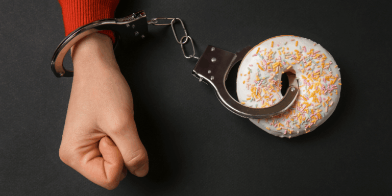 Close-up of a hand in metal handcuffs attached to a frosted donut with colorful sprinkles on a dark background, representing food addiction.