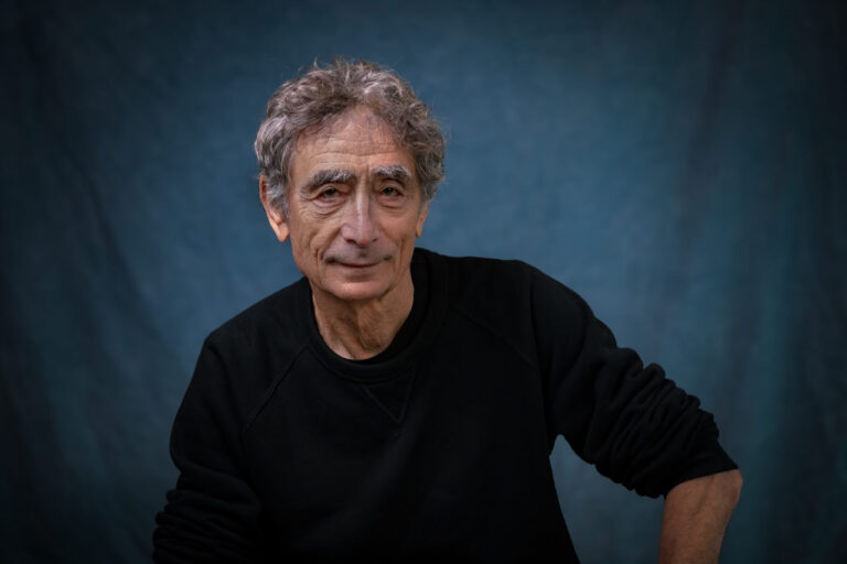 Portrait of Gabor Maté, an elderly man with curly gray hair and bushy eyebrows, wearing a black sweater, smiling subtly while leaning slightly forward on a blue backdrop.