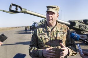 A military general in camouflage uniform with rank insignia speaks into microphones held by reporters, standing near a tank and artillery in an outdoor field, with other troops and equipment visible in the background.