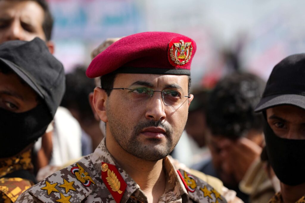 A group of Houthi soldiers in camouflage uniforms with insignia, standing in formation during a military parade, led by a commanding officer.