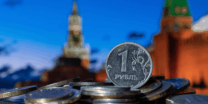 A pile of metallic ruble coins in the foreground, with the Kremlin’s distinctive towers and architecture visible in the background under a clear blue evening sky.