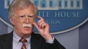 John Bolton, with glasses and a mustache, speaks at a White House press conference podium, wearing a suit and tie, with a government official backdrop.