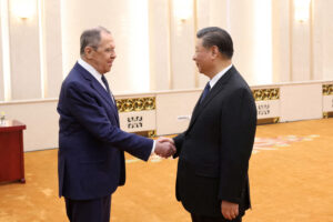 A photo of two men in suits shaking hands in a formal room with ornate decor, identified as Russian Foreign Minister Sergey Lavrov and Chinese President Xi Jinping at the SCO summit, with a desk and patterned carpet in the background.