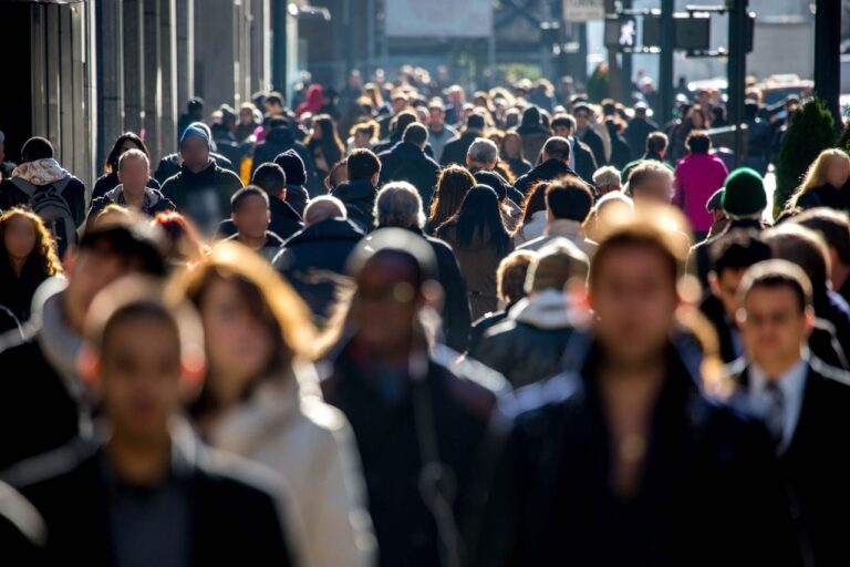 A crowded city street filled with a diverse group of pedestrians walking along the sidewalk, surrounded by tall buildings and traffic in the background.