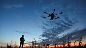 Silhouette of a person standing in a field, controlling a six-rotor drone against a colorful dusk sky with scattered clouds and tall grasses.