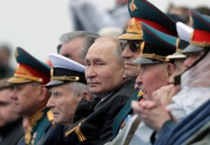 A group of Russian military officials in ornate uniforms, with Vladimir Putin seated centrally among his generals, all gazing forward during a formal event.