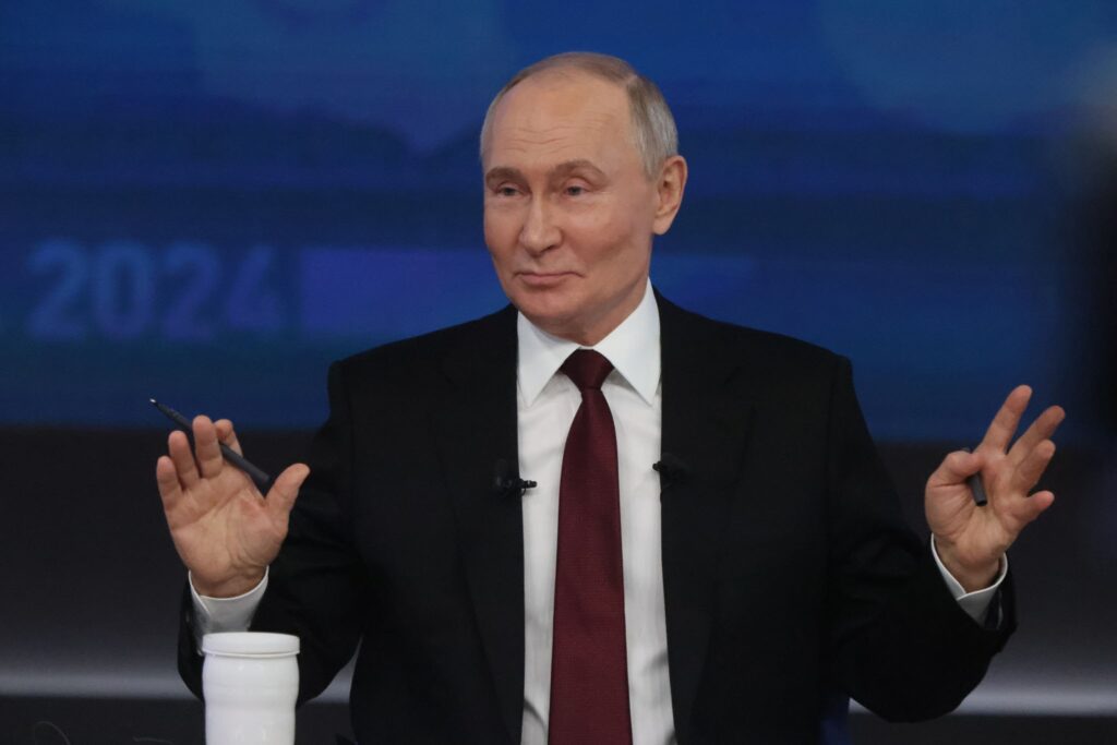 A man in a dark suit and red tie gestures with both hands while holding a pen, speaking at a podium with a blue background displaying the year 2024.