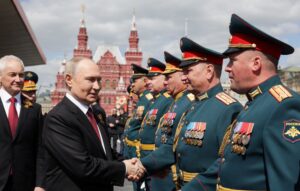 A man in a black suit with a red tie shakes hands with a line of military officers wearing green uniforms with red hats and numerous medals, set against the backdrop of the Kremlin’s distinctive red architecture under a cloudy sky.