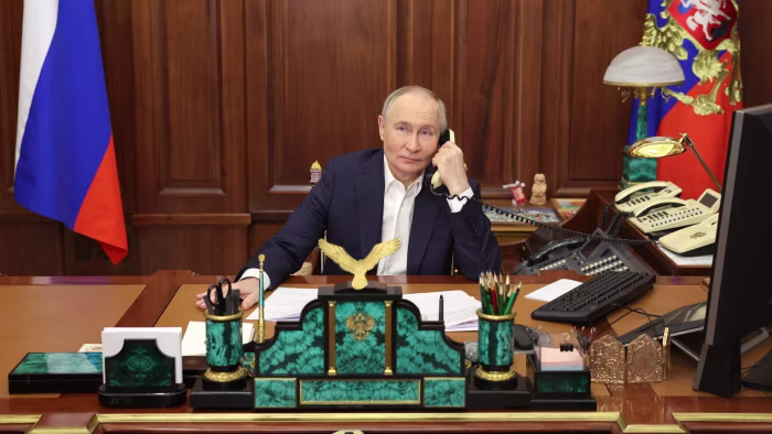 Russian President Vladimir Putin seated at a large wooden desk in his office, holding a telephone receiver to his ear, with the Russian flag to his left, a golden eagle emblem in front, multiple landline phones, a computer monitor, and various office supplies including pens and papers; the room features wooden paneling and a coat of arms in the background.