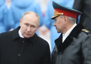 A close-up image of two men in formal attire, one in a black coat and the other in a military uniform with a red and green cap adorned with gold embroidery, standing side by side against a blurred backdrop of blue-uniformed figures.