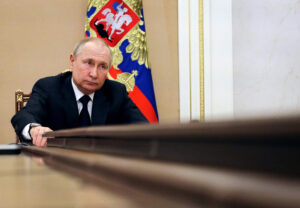 Vladimir Putin in a dark suit and tie, leaning on a large wooden table with a serious expression, with the Russian presidential flag featuring a double-headed eagle in the background.