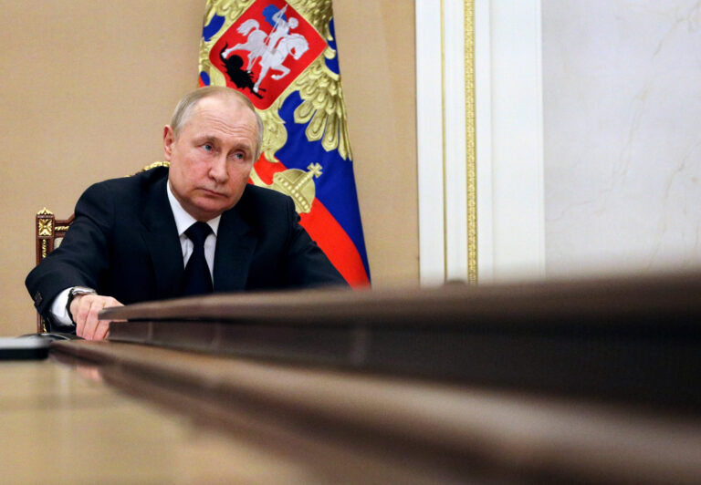 Vladimir Putin in a dark suit and tie, leaning on a large wooden table with a serious expression, with the Russian presidential flag featuring a double-headed eagle in the background.