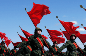 A group of soldiers in green camouflage uniforms with red sashes marching in formation, each holding a red flag against a clear blue sky.
