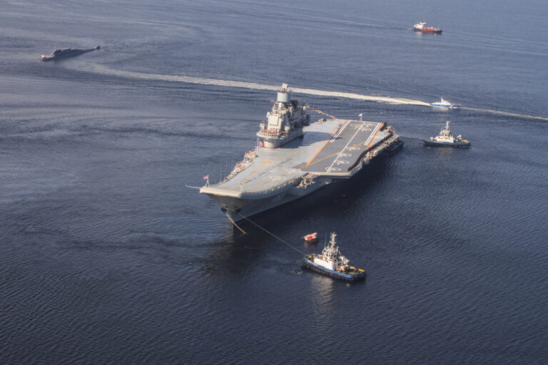 An aerial view of a large aircraft carrier with a flat deck, guided by several tugboats on a calm sea.