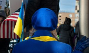 The back of a person with blonde hair, wearing a blue beret, a blue coat, and a yellow scarf. To the left of the person, a partially visible American flag is displayed, and a Ukrainian flag is also present. The background is out of focus but shows what appears to be an outdoor gathering or protest with other people and buildings.