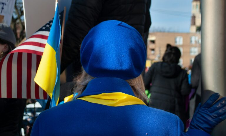 The back of a person with blonde hair, wearing a blue beret, a blue coat, and a yellow scarf. To the left of the person, a partially visible American flag is displayed, and a Ukrainian flag is also present. The background is out of focus but shows what appears to be an outdoor gathering or protest with other people and buildings.