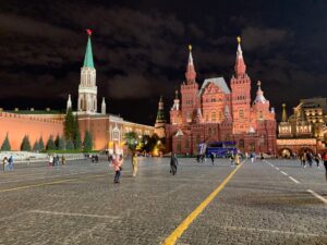 A nighttime view of Red Square in Moscow, featuring the brightly lit Kremlin walls and towers on the left, the ornate red and green domes of Saint Basil's Cathedral on the right, and a scattering of people walking across the cobblestone plaza under a cloudy sky.