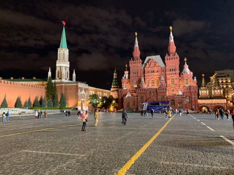A nighttime view of Red Square in Moscow, featuring the brightly lit Kremlin walls and towers on the left, the ornate red and green domes of Saint Basil's Cathedral on the right, and a scattering of people walking across the cobblestone plaza under a cloudy sky.