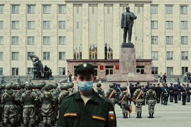 A Russian soldier in a green uniform stands in the foreground, with masked troops and a Soviet statue behind, set against a government building.