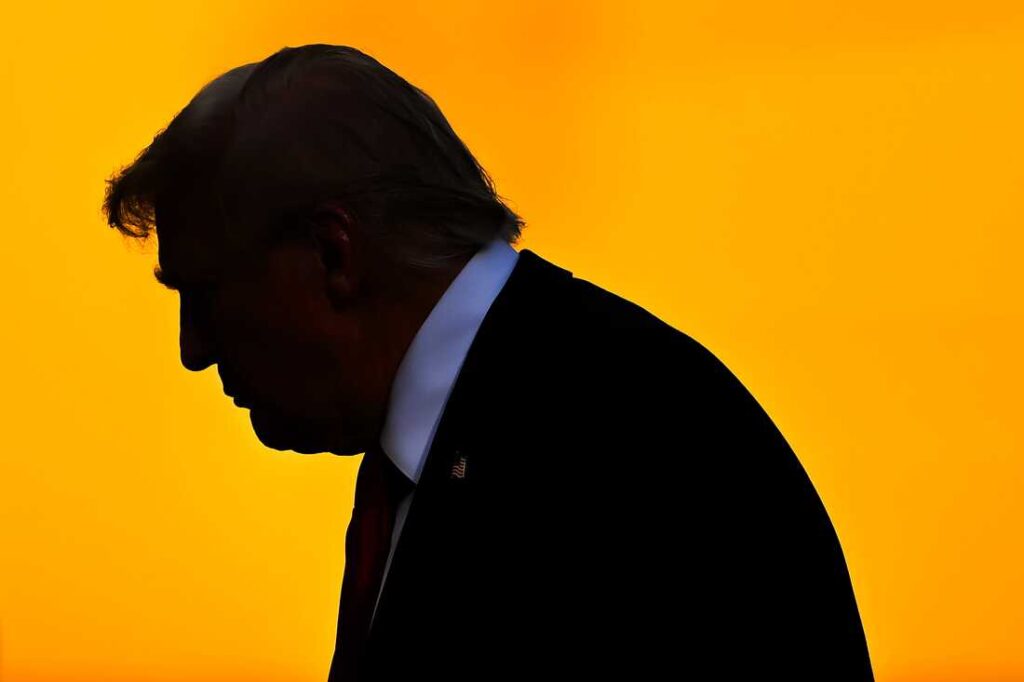 A black silhouette of Donald Trump in a suit and red tie, set against a bright orange background, capturing his profile as he walks, with a pin on his lapel faintly visible.