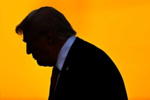 A black silhouette of Donald Trump in a suit and red tie, set against a bright orange background, capturing his profile as he walks, with a pin on his lapel faintly visible.