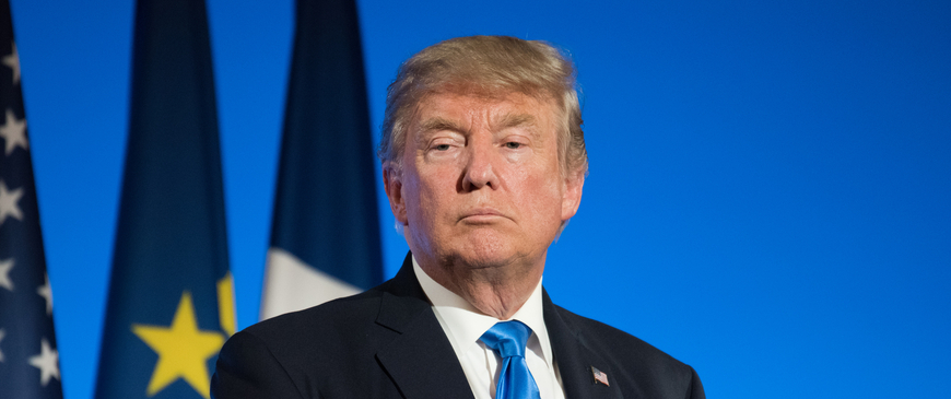 A portrait of Donald Trump in a dark suit and blue tie, standing with a serious expression against a blue background with American and European Union flags.