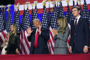 Image of Donald Trump waving at a rally, flanked by family members including a woman in a gray suit, a young man in a blue suit, and others, with numerous American flags in the background.