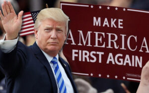 An image of a man with blonde hair in a dark suit and blue tie raising his right hand, standing in front of an American flag, with a large sign reading "Make America First Again" held by a crowd in the background.