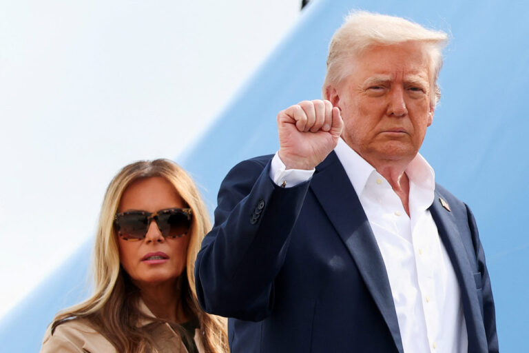 Donald Trump, in a dark suit and white shirt, raises his right fist while standing in front of an airplane. Melania Trump, wearing sunglasses and a beige outfit, stands slightly behind him with a neutral expression.