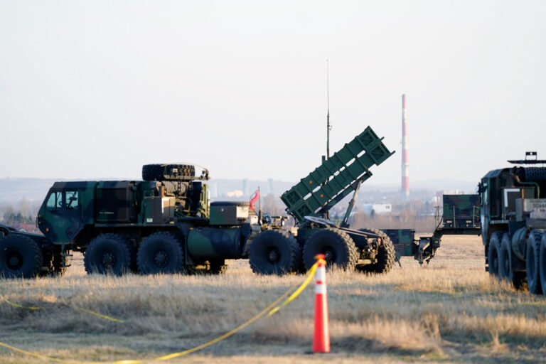 A military rocket launcher system with multiple wheels, angled upward, positioned in a grassy field with yellow caution tape and an orange cone in the foreground. Another similar vehicle is partially visible to the right, and a factory smokestack looms in the background under a pale sky.