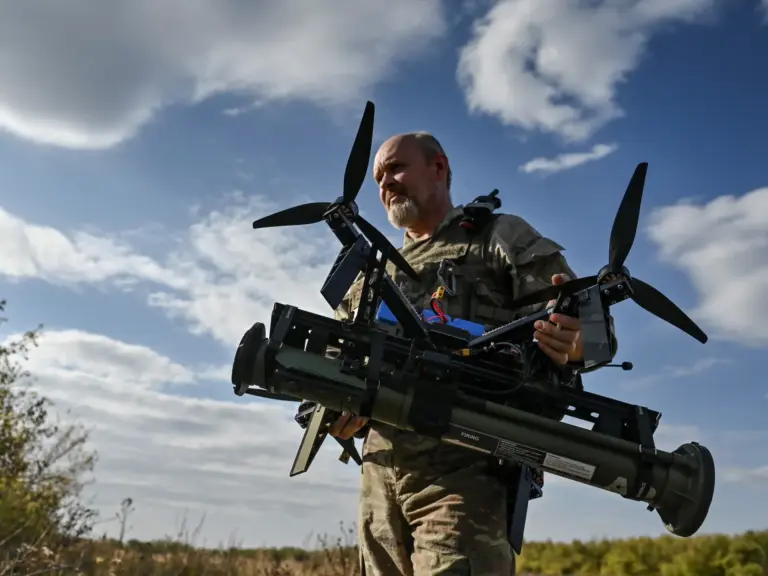 A Ukrainian soldier in camouflage holds a drone with attached missile components against a backdrop of a cloudy sky and open field, symbolizing the country’s advanced drone warfare tactics.