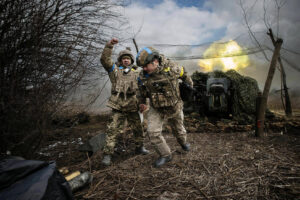 Two Ukrainian soldiers in camouflage gear, one raising a fist in triumph, stand near a camouflaged military vehicle as an explosion lights up the sky. Bare trees and scattered debris surround the scene.