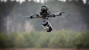 A military drone carrying a cylindrical explosive payload flies over a forested area, with its propellers spinning and a thin wire trailing beneath.