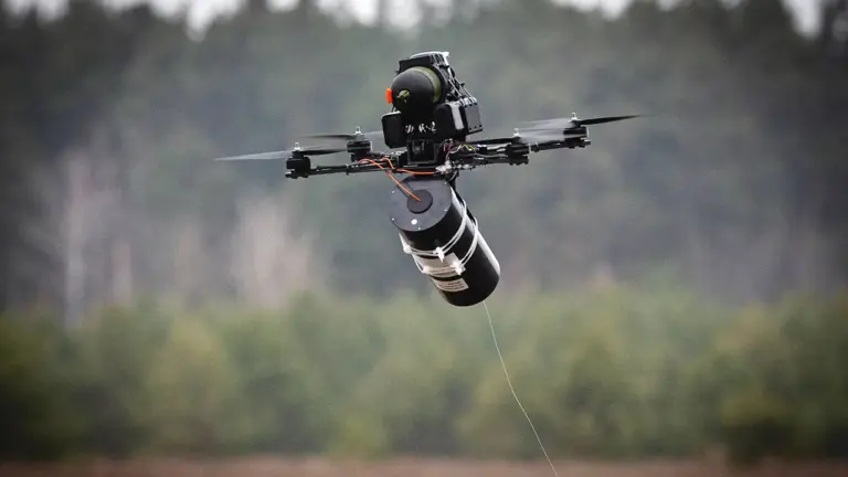 A military drone carrying a cylindrical explosive payload flies over a forested area, with its propellers spinning and a thin wire trailing beneath.