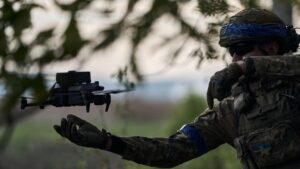 A Ukrainian soldier in camouflage gear with a blue armband and a Ukrainian flag patch launches a drone with an outstretched hand, surrounded by a blurred forest background.