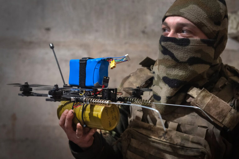 A camouflaged Ukrainian soldier holding a modified drone equipped with a yellow explosive canister and a blue battery pack, standing against a rough concrete wall, highlighting the ingenuity of Ukraine's drone warfare strategy.